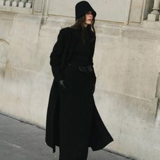 Lena wears a black hat, black long coat, black maxi dress, black leather belt, and black gloves.