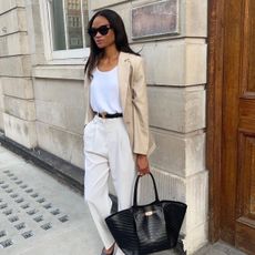 One woman wears a beige blazer, white tank top, black belt, ivory trousers and black tote bag. 