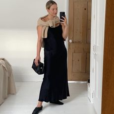 Influencer Alexis Foreman posing for a mirror selfie wearing a black slip dress with a neutral sweater tied around her shoulders