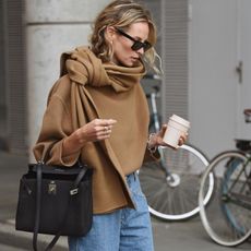 female fashion influencer Anouk Yve with wavy blonde hair wearing black sunglasses, a camel tan scarf sweater, coffee in hand, black Hermes bag on the arm, and high-waisted jeans