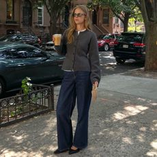 Scandi style influencer and creative Clara Dyrhauge poses on a New York City sidewalk holding a cup of coffee wearing a fitted gray cardigan sweater, relaxed dark-wash jeans, and black heels