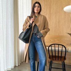 British fashion influencer Anna Howard poses in her chic wood-paneled London kitchen wearing a camel coat, gray sweater, black tote bag, black belt, and straight-leg jeans