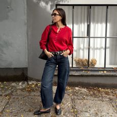 British style influencer Jessica Skye poses in front of a building wearing black oval sunglasses, a red button collar henley sweater, Khaite studded belt, and dark wash barrel jeans, and black ballet flats