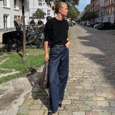 Art director and fashion influencer Clara Dyrhauge on the street in Europe wearing dark denim jeans, a black belt, a black short-sleeve knit top, black pointed-toe Aeyde shoes, and carrying a woven handbag.